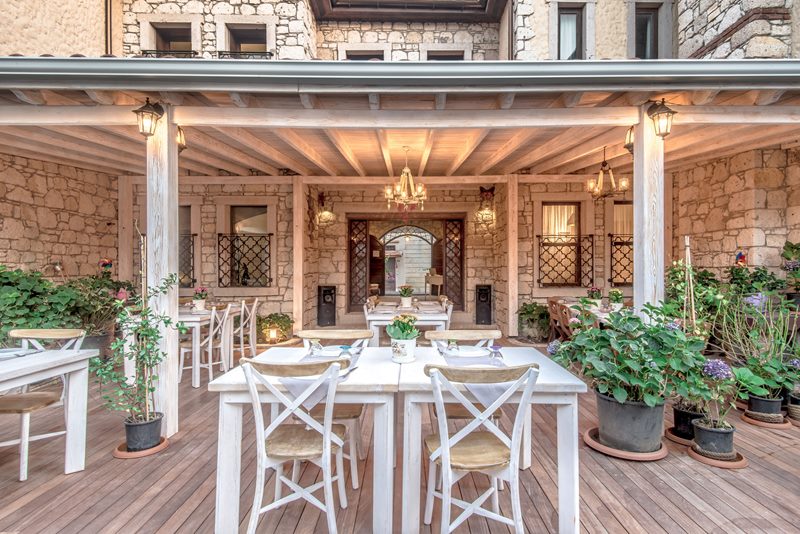 D'Core Alaçatı Hotel ahşap pergolalı terasında beyaz yemek masaları, avize ve saksı çiçekleri.