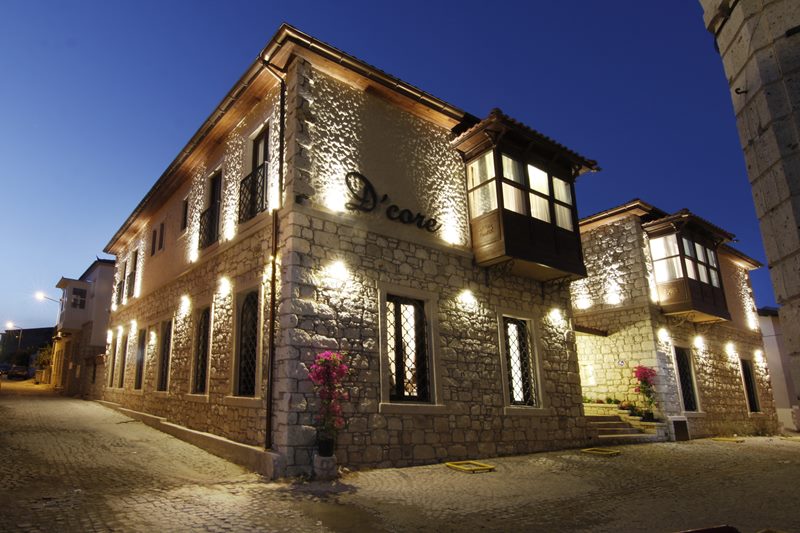 D'Core Alaçatı Hotel'in gece ışıklandırmalı taş cephesi, ahşap cumbası, begonviller ve otelin sokak girişi.