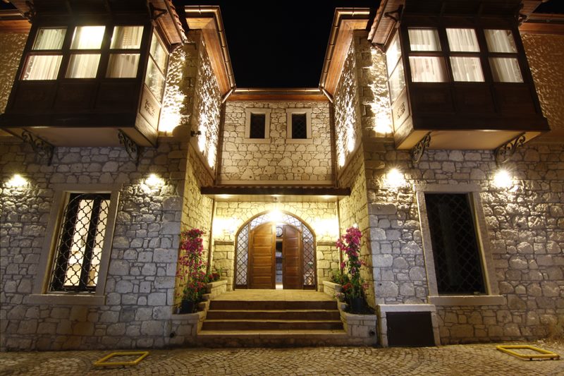 D'Core Alaçatı Hotel'in kemerli ahşap giriş kapısı, basamaklar, pembe begonviller ve cumbanın gece görünümü.