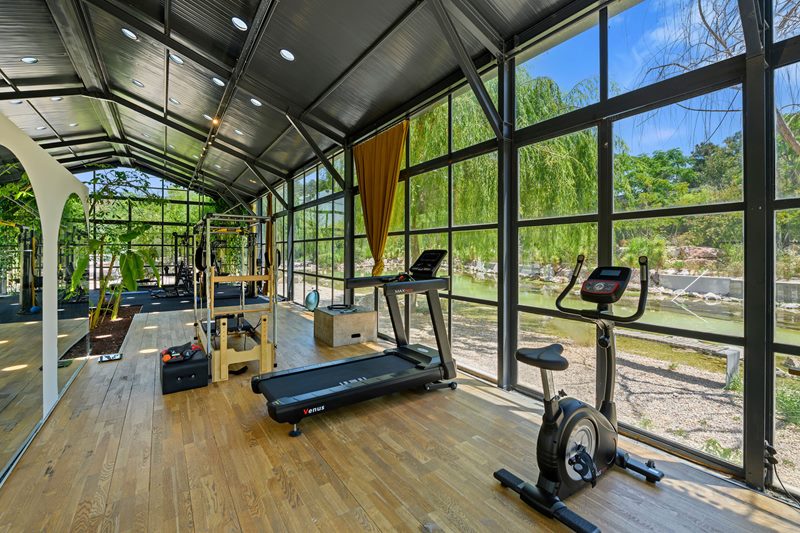Urla'daki lüks bir villanın nehir manzaralı, geniş cam cepheli ve spor aletleriyle donatılmış ferah spor salonunun mimari fotoğrafı.