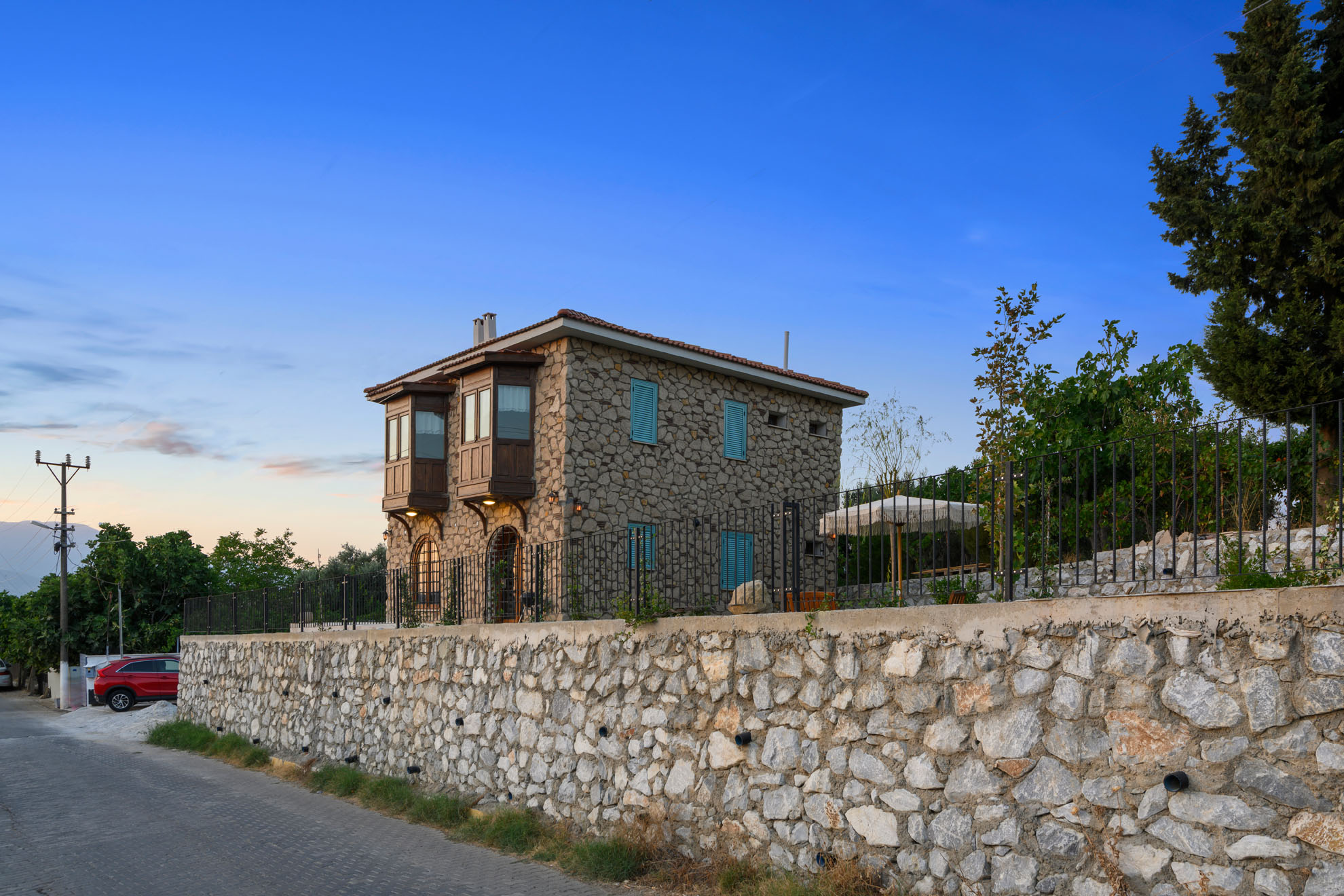 Gün batımı ışığıyla aydınlanan mavi panjurlu ve geleneksel mimarili İzmir Torbalı taş ev projesinin dış mekan fotoğrafı.