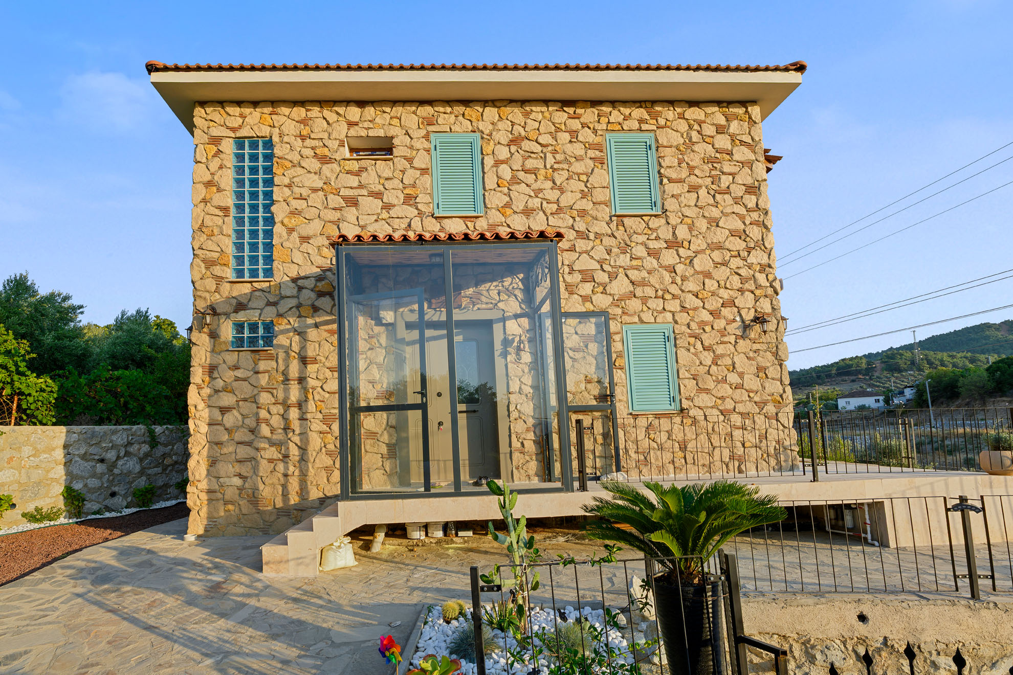 İzmir Torbalı'daki doğal taş ev, altın saatte turkuaz panjurları ve modern cam girişiyle huzurlu bir emeklilik yaşamını yansıtıyor.