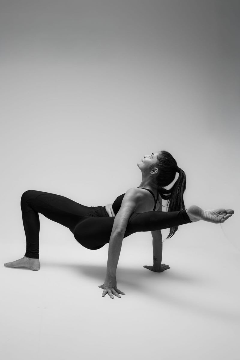 İzmir Menemen'deki stüdyoda açık gri fon önünde zorlu bir asana pozu sergileyen yoga eğitmeni Melis'in siyah beyaz yoga fotoğrafı.