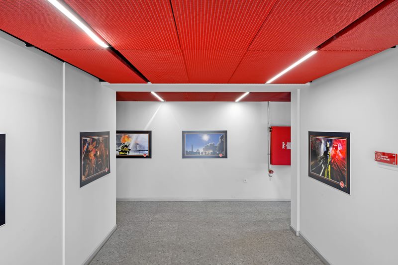 İtfaiye istasyonu holünde kırmızı metal asma tavan kasası, lineer LED'ler ve duvarlarda çerçeveli fotoğraflar.