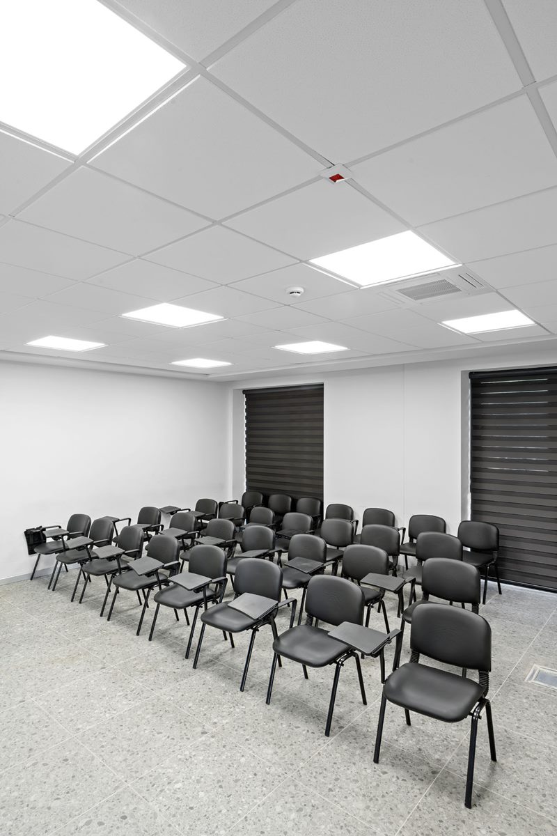 İtfaiye eğitim salonunda modüler tavan ve LED paneller altında sandalye sıraları ve koyu zebra perdeler.