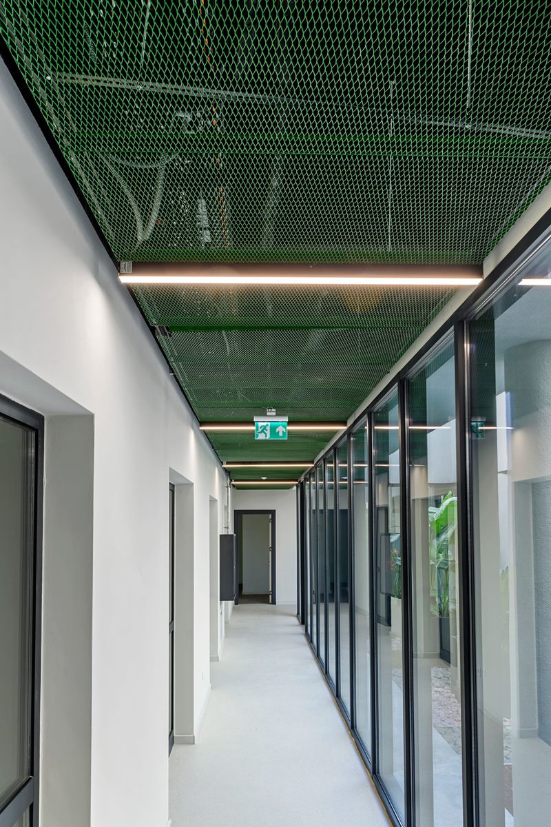 Yeşil metal tavanlı anaokulu koridorunda boydan boya lineer LED'ler ve iç bahçeye bakan cam cephe, dikey kadraj.