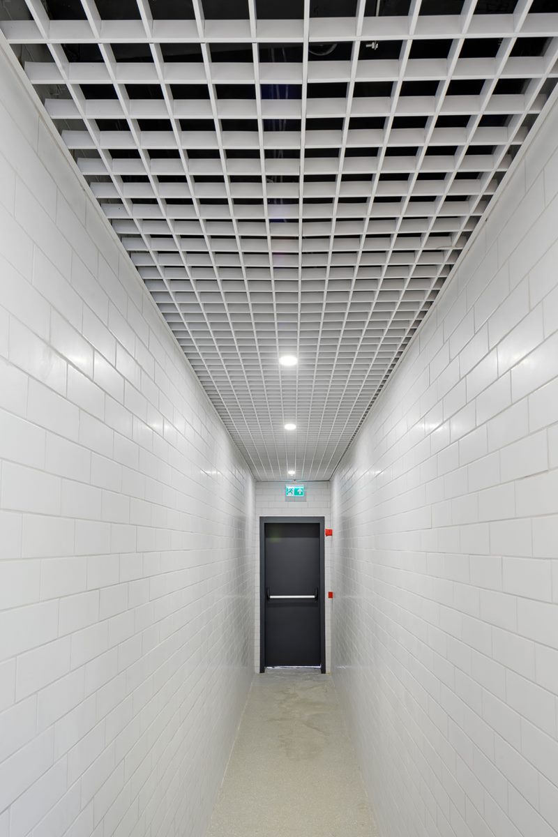 Anaokulu servis koridorunda hücre tipi (grid) metal asma tavan, spot aydınlatma ve beyaz fayanslı duvarlar.