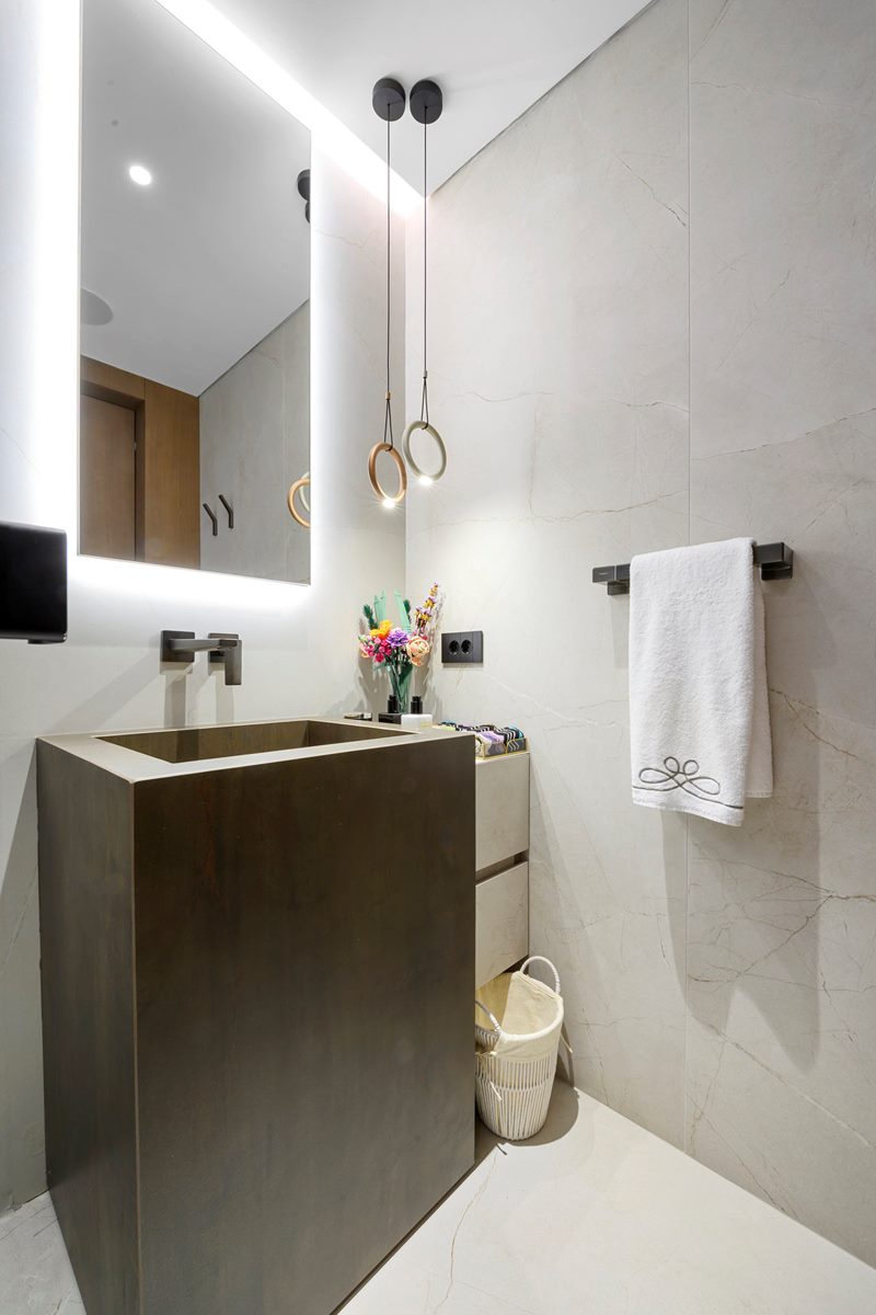 Banyoda koyu ahşap lavabo dolabı, aydınlatmalı ayna ve gri taş kaplama duvarlar, özel üretim banyo mobilyası.
