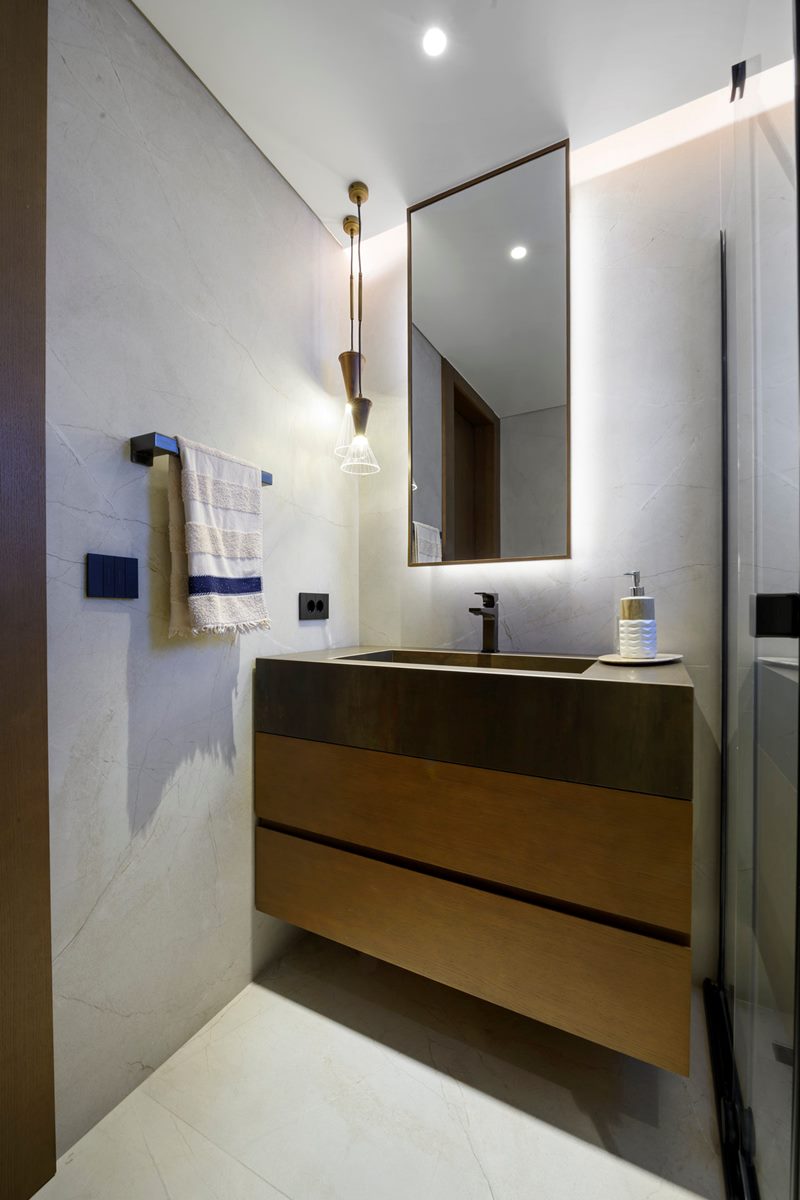 Dikey kadrajda banyonun ahşap lavabo ünitesi, arkadan aydınlatmalı ayna ve spot ışıkları, villa iç mekân fotoğrafı.