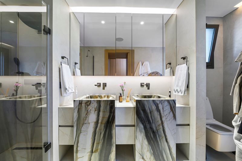 Ebeveyn banyosunda yeşil damarlı mermer çift lavabo, aydınlatmalı ayna dolabı ve cam duşakabin, villa iç mekân.
