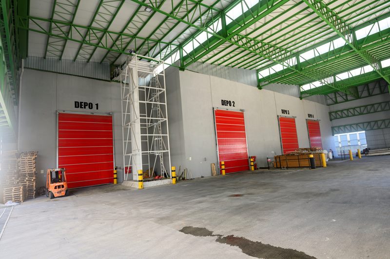 Yüksek tavanlı antrepo koridorunda Depo 1, 2 ve 3'ün kırmızı hızlı kapıları, çelik merdiven ve beton zemin.