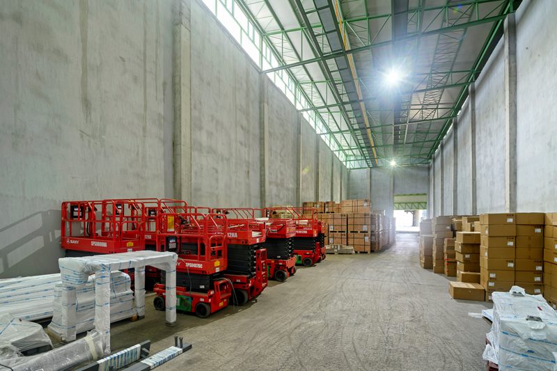 Antrepo deposunda sıralanmış kırmızı makaslı iş platformları, paletli yükler ve yüksek tavan, geniş açı.