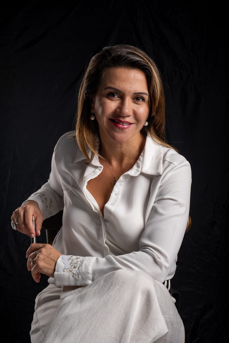 Dilara Karaçoban kişisel marka fotoğraf çekimi koyu fonda beyaz gömlek ve pantolonla sandalyede dolma kalem tutan renkli stüdyo portresi.
