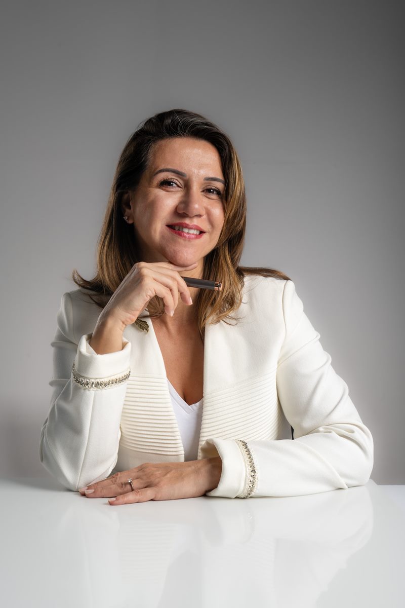İzmir Dilara Karaçoban kurumsal portre fotoğraf çekimi açık fonda sağ elinde dolma kalemle masada tebessüm eden renkli portresi.
