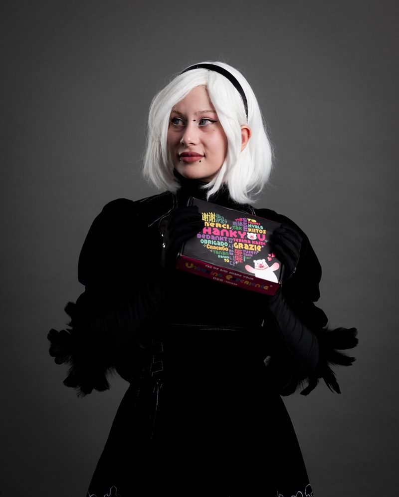 Stüdyoda gri fon önünde elinde kutu tutan beyaz peruklu 2B kostümlü modelin dikey cosplay fotoğraf çekimi karesi.
