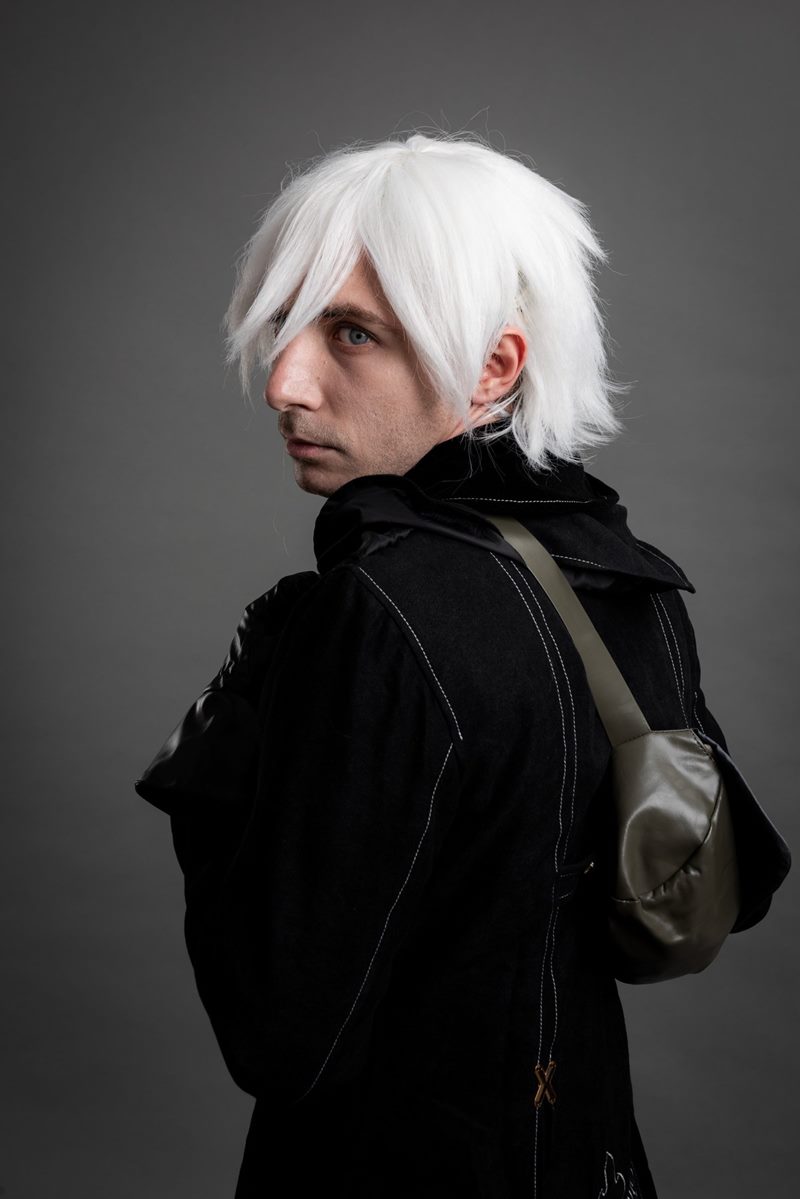 Stüdyoda gri kağıt fon önünde omuz üzerinden geriye bakan sırt çantalı NieR Automata 9S cosplay fotoğraf çekimi.