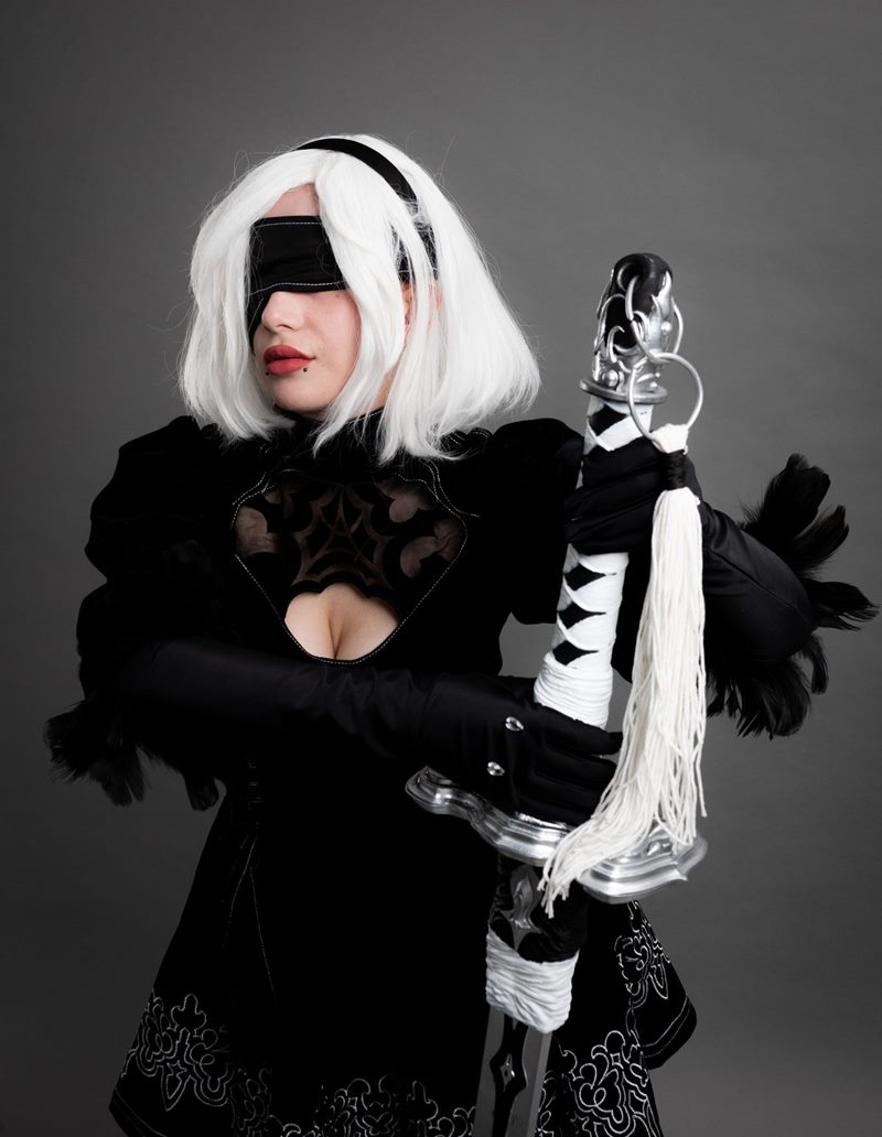 Stüdyoda gri kağıt fonda önünde dikey tuttuğu kılıcın kabzasını kavrayan göz bantlı 2B cosplay fotoğraf çekimi.