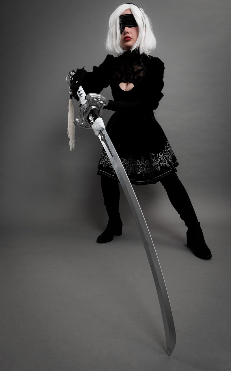 Gri stüdyo fonunda uzun kılıcını ön plana doğru uzatarak güçlü bir duruş sergileyen 2B cosplay fotoğraf çekimi.