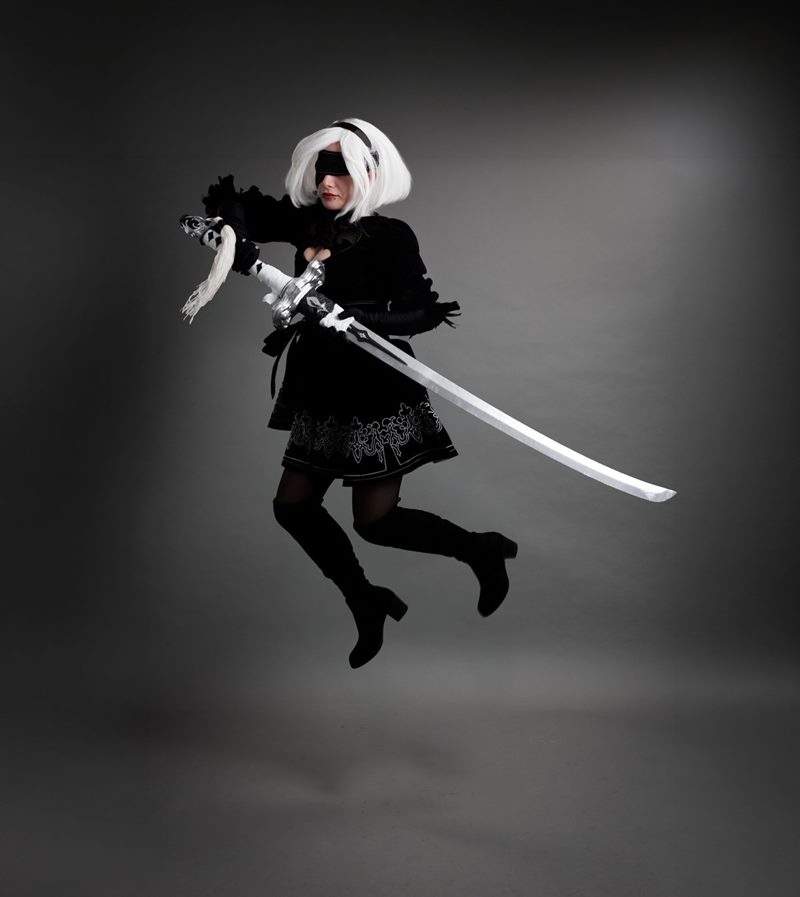 Gri kağıt fon önünde elinde uzun kılıcıyla havaya sıçrayarak aksiyon pozu veren 2B İzmir cosplay fotoğraf çekimi.