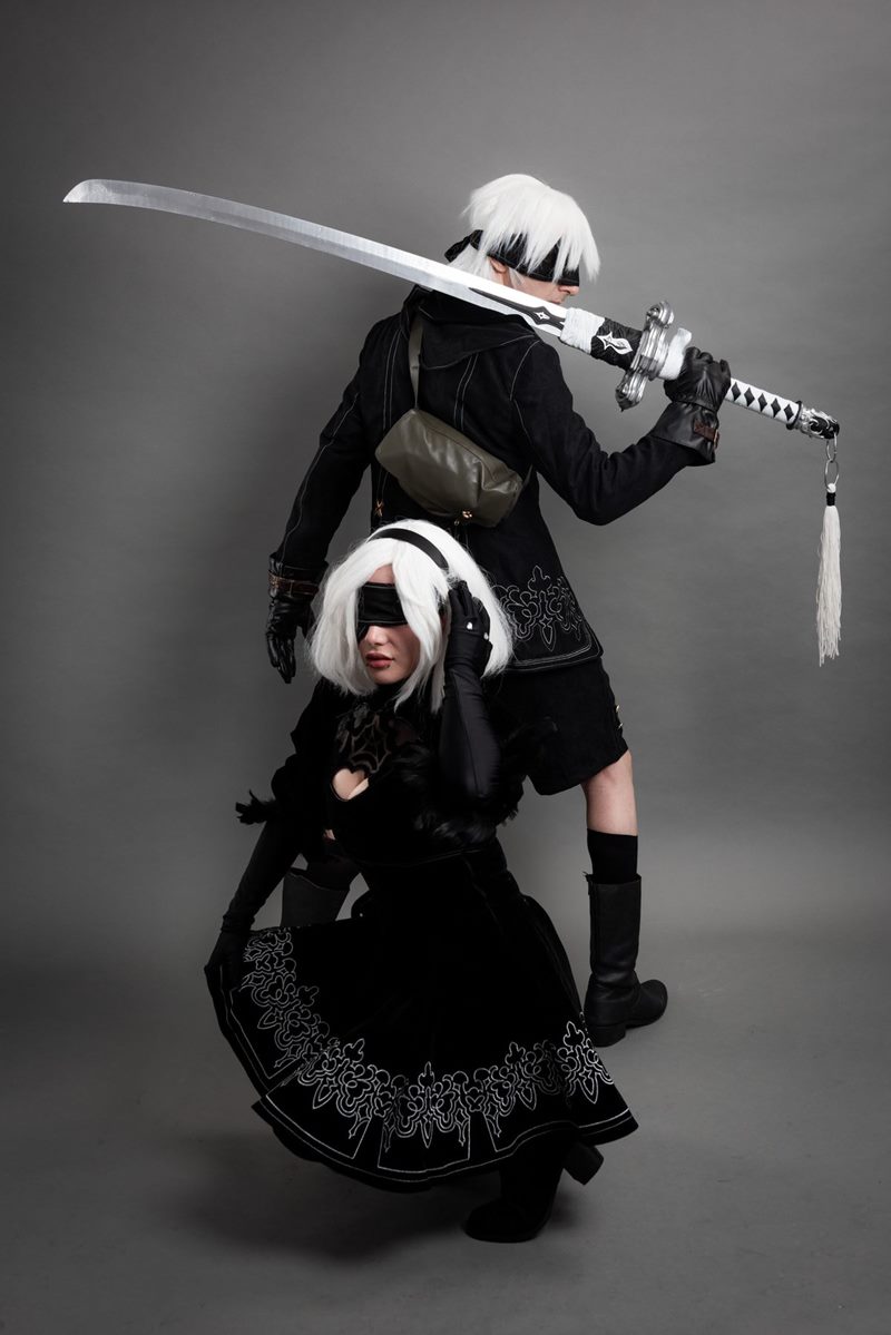 İzmir cosplay fotoğraf çekimi kapsamında gri fonda çömelmiş 2B ve arkasında uzun kılıç tutan 9S ikili stüdyo pozu.