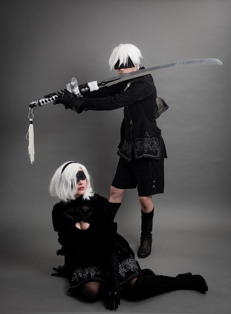 Gri stüdyo fonunda yerde oturan 2B ve arkasında kılıcını yatay uzatan 9S karakterleri cosplay fotoğraf çekimi.