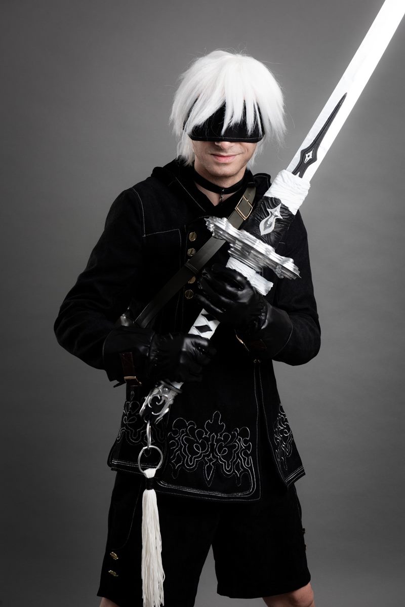 İzmir cosplay fotoğraf çekimi stüdyosunda gri fonda kılıcını göğsünde çapraz tutan göz bantlı 9S portre karesi.