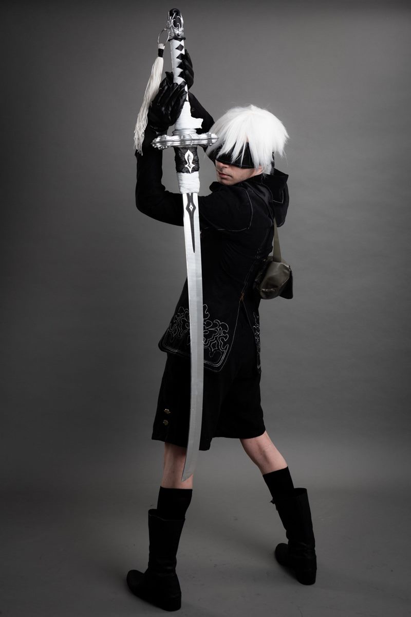 Gri kağıt fonda el yapımı uzun kılıcını dikey tutarak omuz üstünden bakan 9S karakteri İzmir cosplay fotoğraf çekimi.