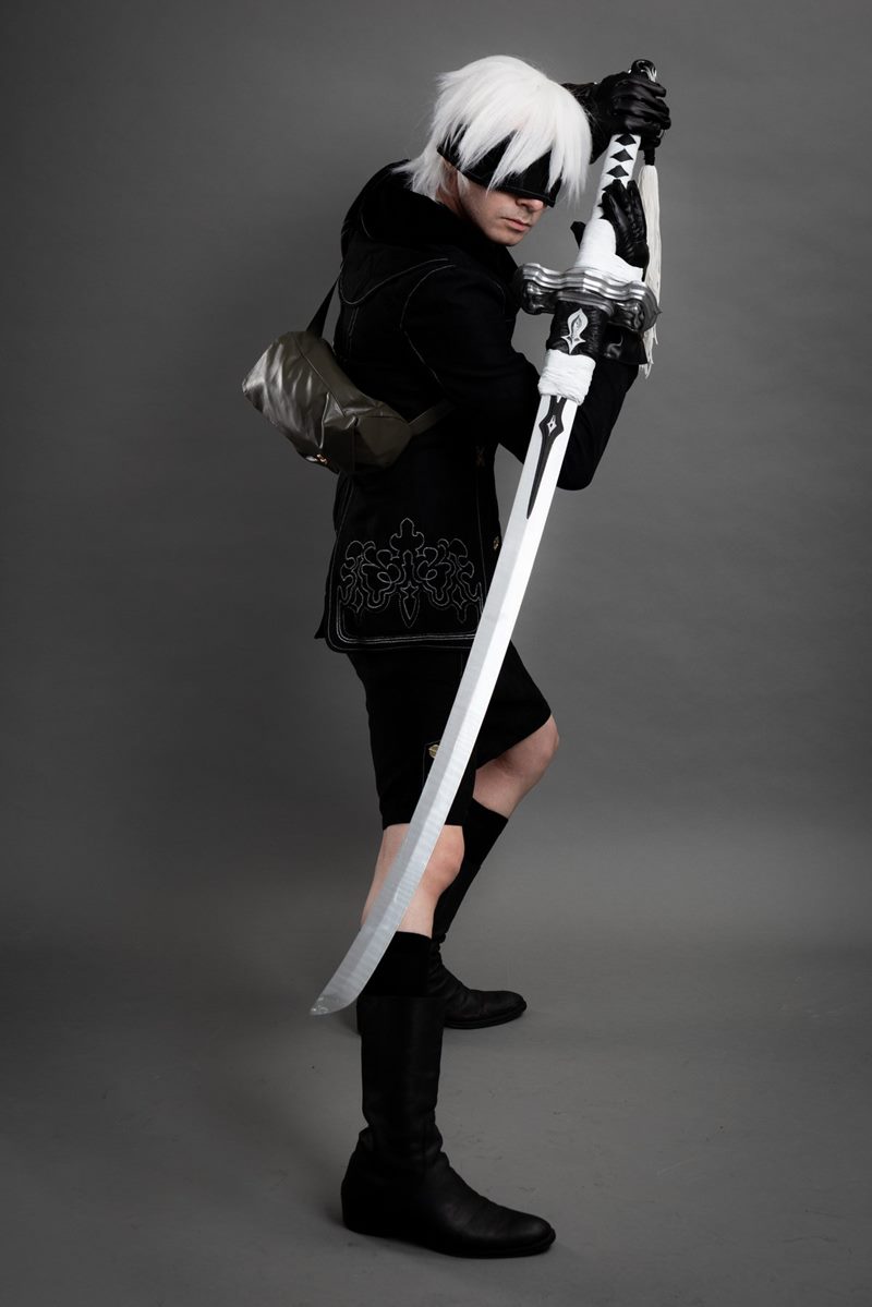 Stüdyoda gri kağıt fon önünde yan profilden uzun kılıcını tutarak poz veren NieR Automata 9S cosplay fotoğraf çekimi.