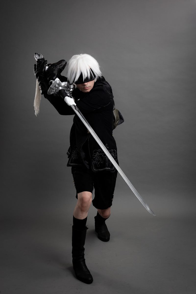 İzmir cosplay fotoğraf çekimi kapsamında gri fonda uzun kılıcını çapraz açıyla tutan göz bantlı 9S tam boy pozu.