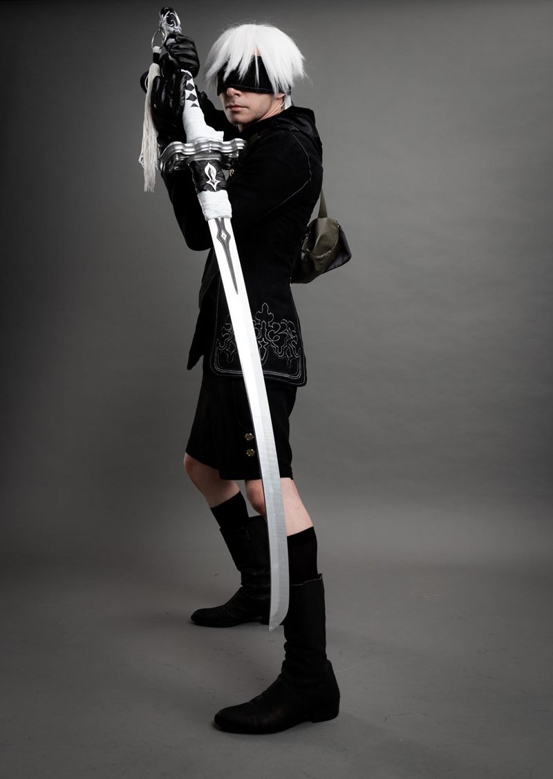 Gri kağıt fon önünde el yapımı kılıcını dikey tutarak kameraya bakan 9S karakteri İzmir cosplay fotoğraf çekimi.