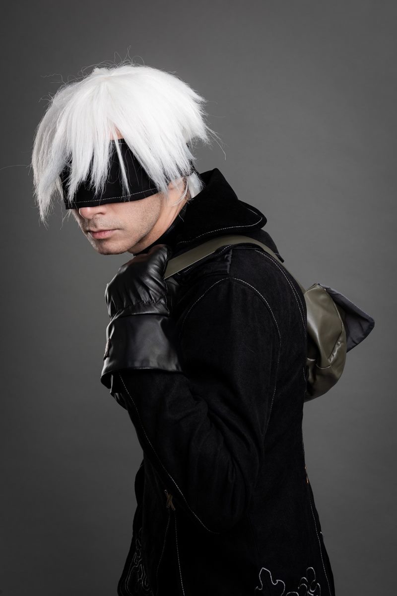 Stüdyoda gri fon önünde deri eldiveniyle çanta askısını tutan göz bantlı 9S yakın plan cosplay fotoğraf çekimi.