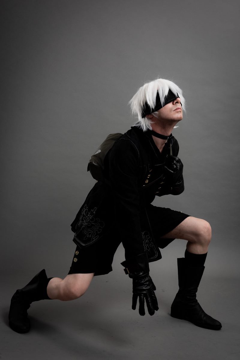 Gri kağıt fonda tek dizini yere koyup başını yukarı kaldıran göz bantlı 9S karakteri İzmir cosplay fotoğraf çekimi.