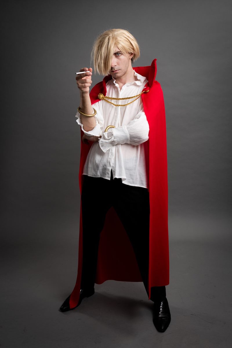 Gri stüdyo fonunda sarı peruklu, kırmızı pelerinli ve beyaz gömlekli kostümüyle tam boy cosplay fotoğraf çekimi.