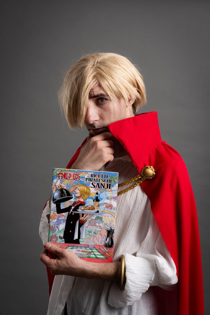 Stüdyoda gri kağıt fon önünde elinde resimli kitap tutan sarı peruklu ve kırmızı pelerinli cosplay fotoğraf çekimi.