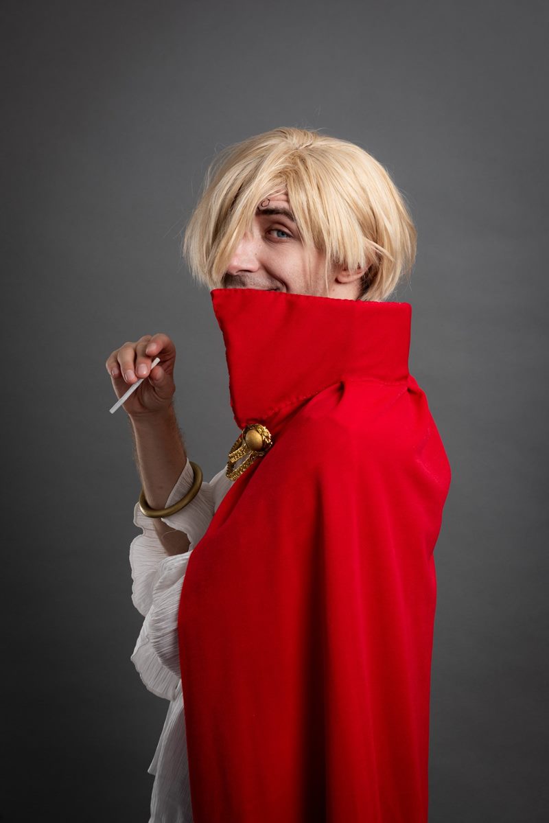 Gri kağıt fonda kırmızı pelerininin dik yakası arkasından kameraya bakan sarı peruklu model cosplay fotoğraf çekimi.