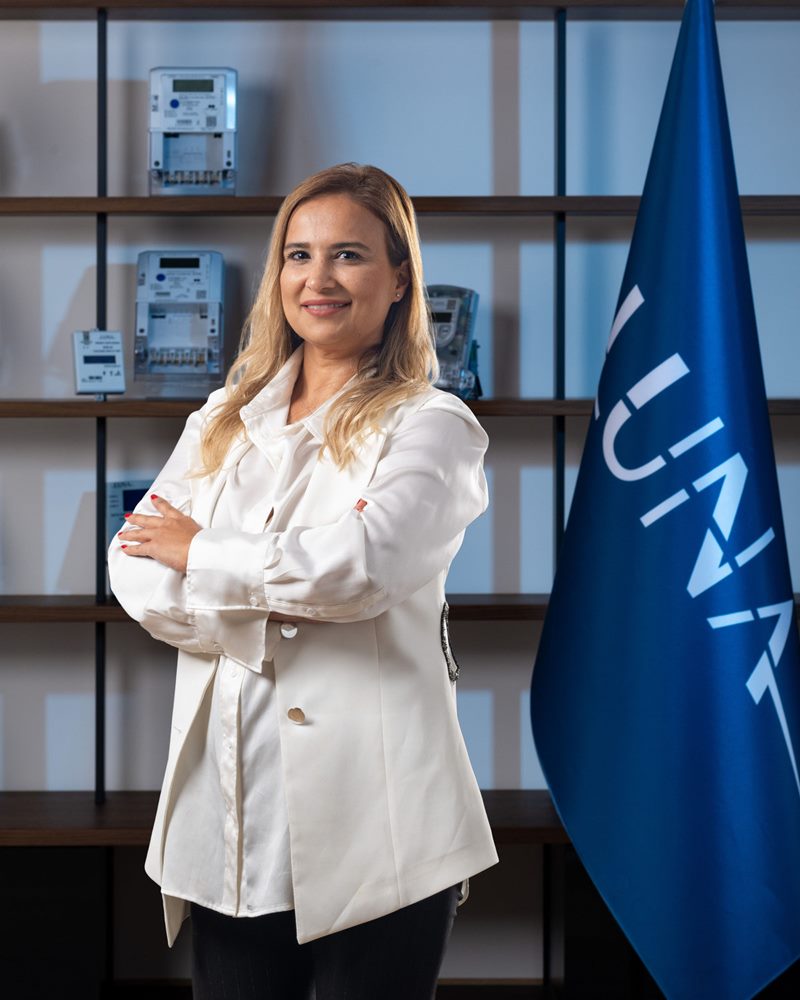 Beyaz ceket ve gömlekle kollarını kavuşturarak gülümseyen sarı saçlı kadının sayaç rafı ve mavi bayrak önündeki dikey portresi.