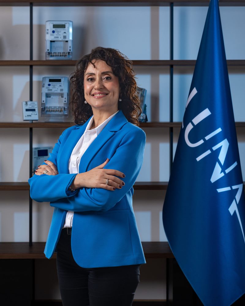 Mavi ceket ve beyaz gömlekle ürün rafları önünde gülümseyen dalgalı saçlı kadın, İzmir kurumsal portre fotoğraf çekimi dikey karesi.
