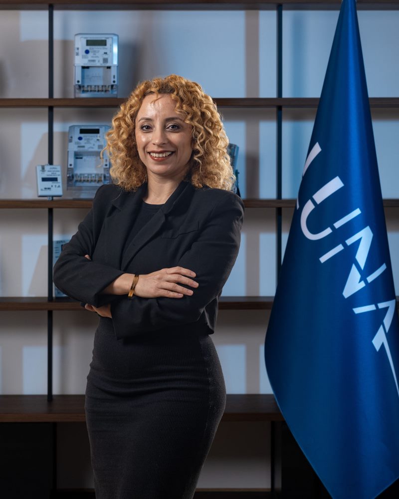 Sarı kıvırcık saçlı ve siyah ceket takımlı kadının mavi aydınlatmalı sayaç rafı ve bayrak önünde kollarını kavuşturarak gülümseyen portresi.