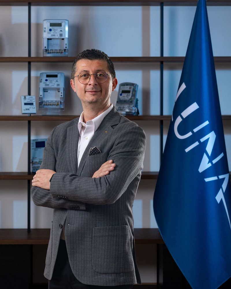 İnce çerçeveli gözlüklü, cep mendilli gri ceket ve beyaz gömlek giyen erkeğin elektronik sayaç rafları ve mavi bayrak önündeki dikey portresi.