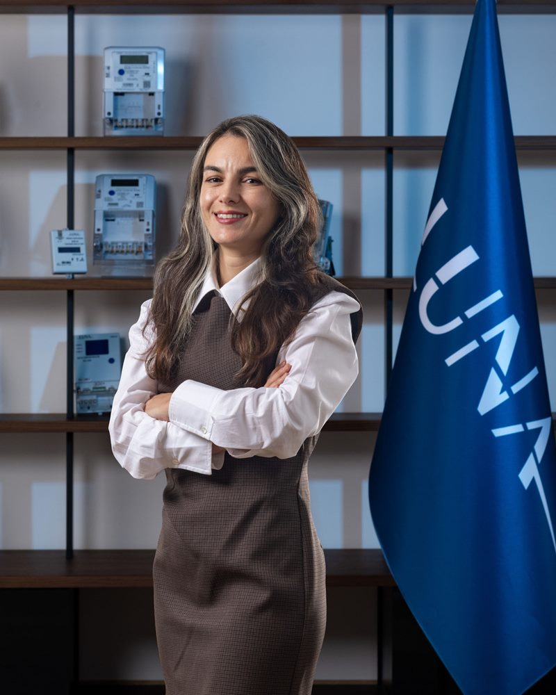 Kahverengi kolsuz elbise ve beyaz gömlekle ürün rafı ve mavi bayrak önünde gülümseyen kadın, İzmir kurumsal portre fotoğraf çekimi.