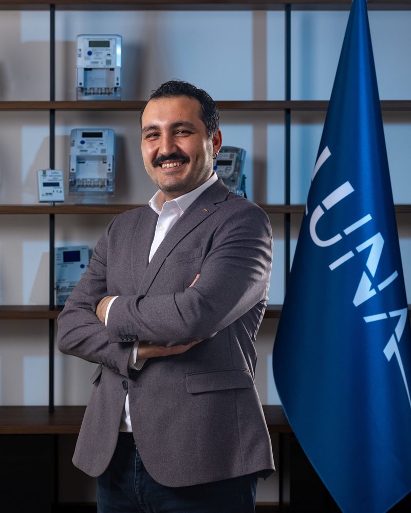 Bıyıklı, gri ceket ve beyaz gömlekli erkeğin elektronik sayaçların dizildiği ahşap raf ve mavi bayrak önünde gülümseyen dikey kadraj portresi.