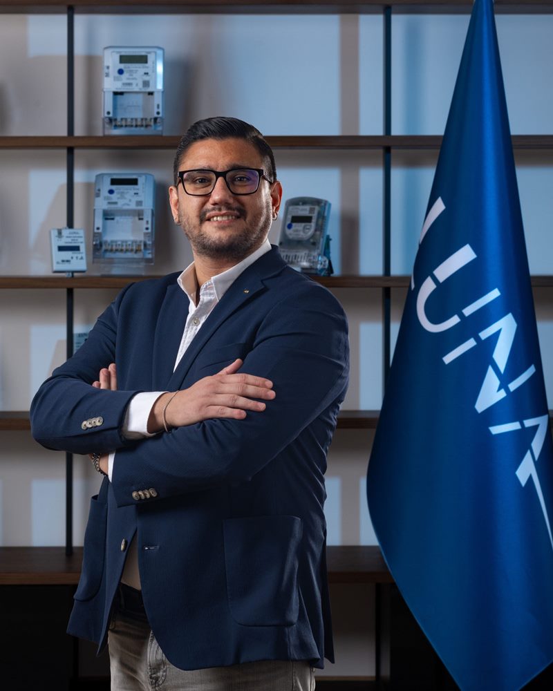 Kalın siyah çerçeveli gözlüklü ve lacivert dokulu ceketli erkeğin mavi aydınlatmalı sayaç rafı ile bayrak önünde gülümseyen dikey portresi.