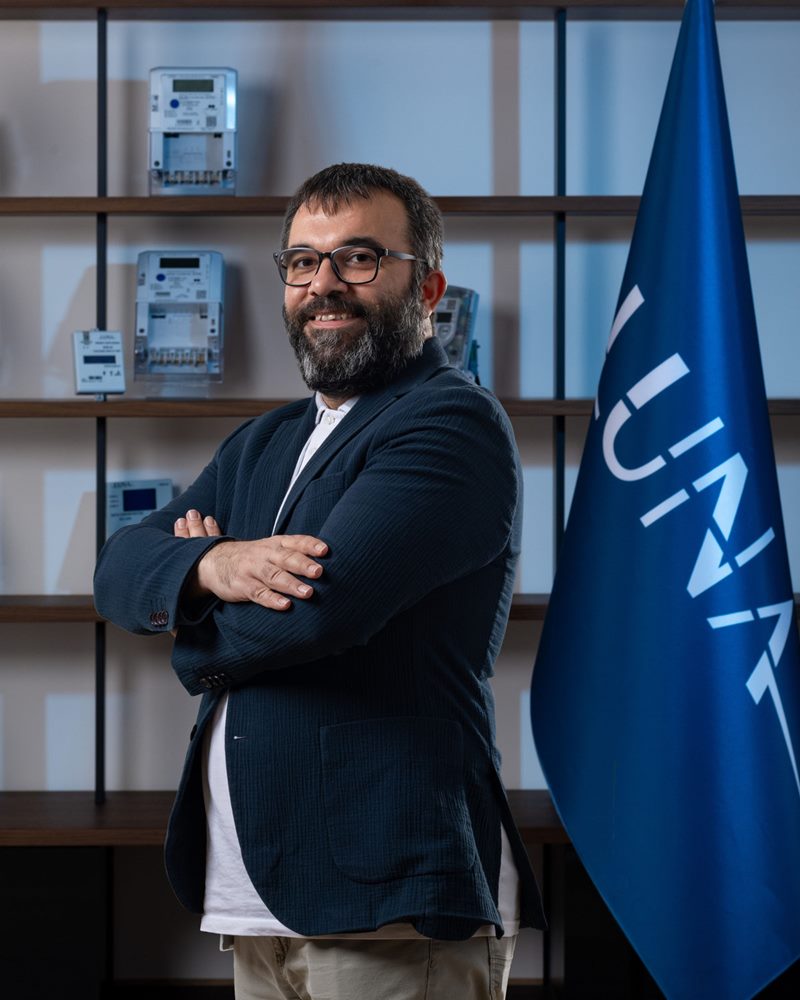 Sakallı, siyah çerçeveli gözlüklü ve lacivert dokulu ceketli erkeğin mavi ışık tonlu sayaç rafları ve bayrak önünde gülümseyen dikey portresi.