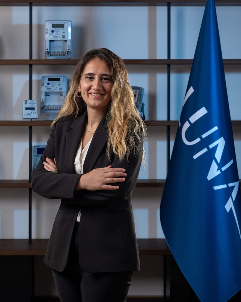Krem rengi kruvaze bluz ve siyah ceketli dalgalı saçlı kadının mavi ışıklı sayaç rafı ve bayrak önünde kollarını kavuşturarak gülümseyen pozu.