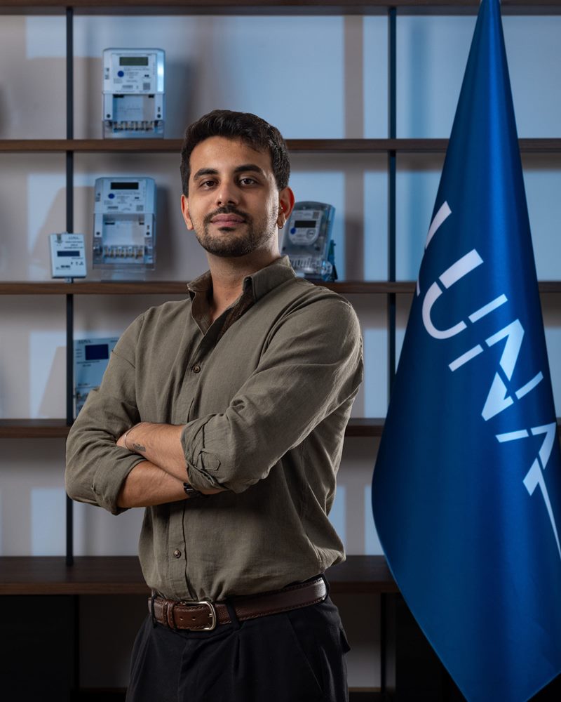 Haki uzun kollu gömlek ve siyah pantolon giyen sakallı erkeğin İzmir'de elektronik sayaç dizili raf ve mavi bayrak önünde çekilen portresi.